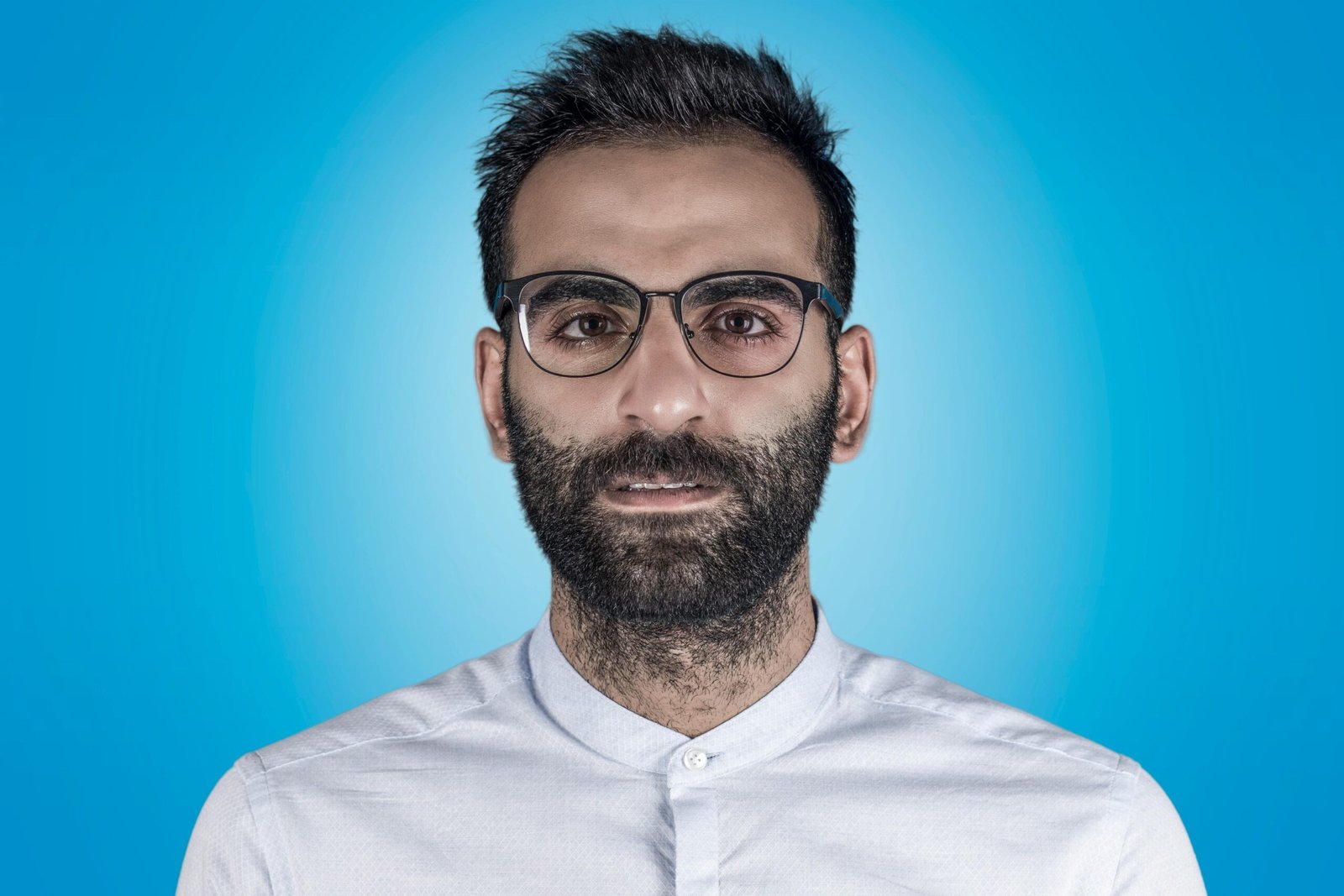 Close-up portrait of a bearded man wearing eyeglasses against a blue backdrop.