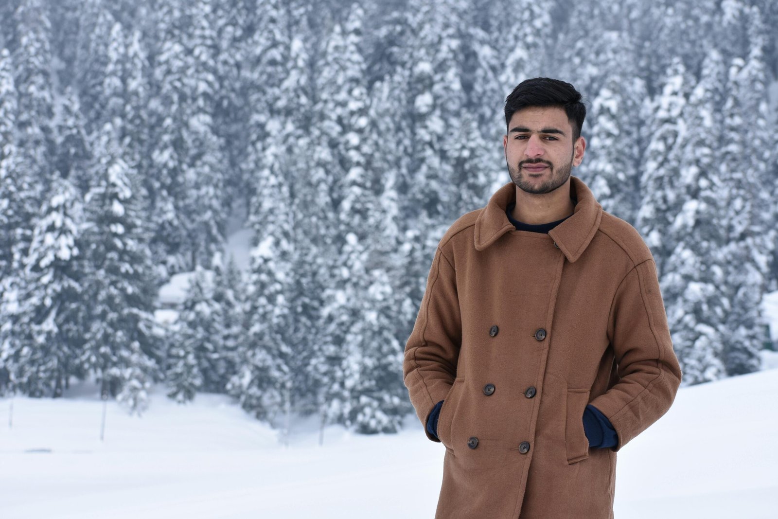 Young man in a brown coat stands in a snowy forest, showcasing winter fashion.